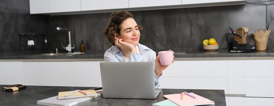 Image of young woman drinks cup of coffee and works from home. Girl browsing website, shops online on laptop and enjoys tea at home.