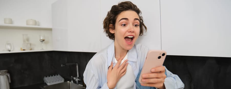 Excited young woman looking amazed at mobile phone screen, chats with someone, sees amazing news on smartphone app, reacts impressed at smth, sitting in kitchen at home.