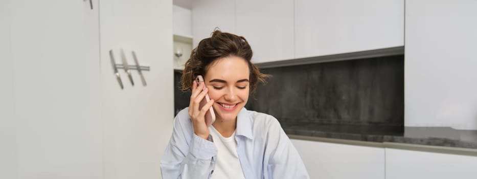 Portrait of smiling young woman talking on mobile phone, laughing, looking down, sitting at home in kitchen.
