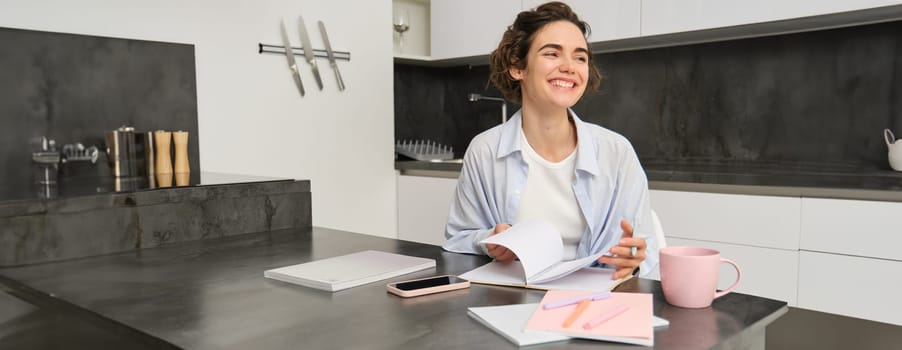 Happy young woman, student doing homework at home, writing in her journal, smiling and looking happy.