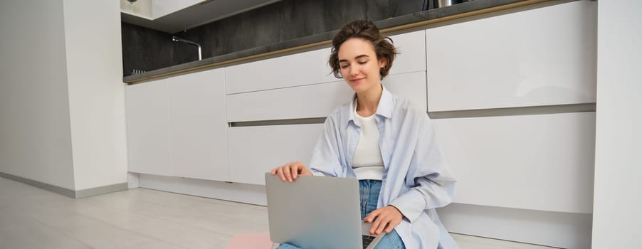 Young woman works from home on kitchen floor, does homework on laptop, using internet on computer, freelancing.