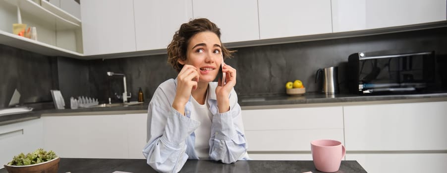 Portrait of woman thinking while talking on mobile phone, sitting with smartphone in kitchen at home and biting pencil with confused face.
