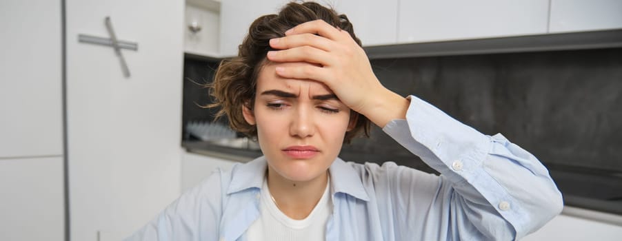 Portrait of woman with headache, sits in kitchen, touches head and grimaces, has painful migraine. Copy space