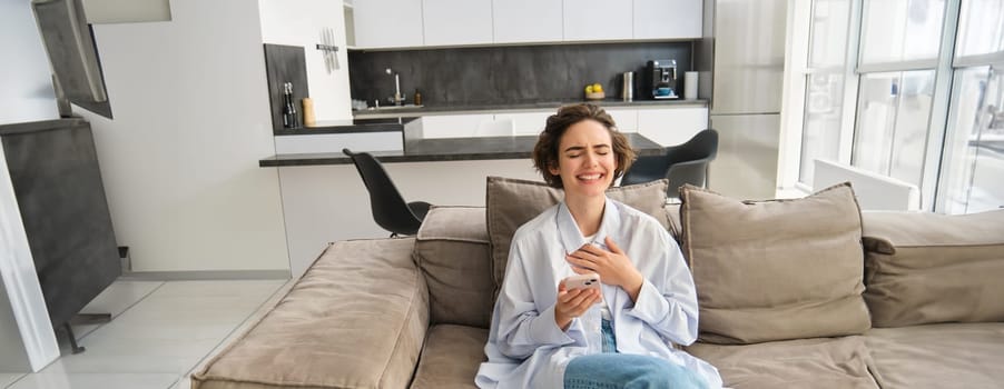 Portrait of happy woman laughing over funny video on phone, holding smartphone, sitting on sofa at home, enjoying scrolling her app feed.