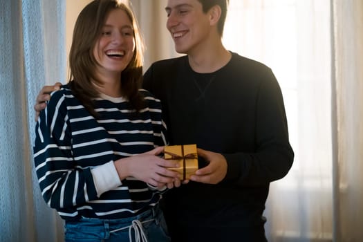 Young man and woman laugh and have a good time together in their cozy home, feel unity. Couple celebrating holidays, valentine's day, anniversary, new year or birthday and give each other cute gifts.