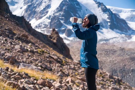 Young sports girl stands on the backdrop of a stunning view with snow-capped mountains and drinks clear water from a bottle. Woman is trail running, training, climbing and drinking isotonic.