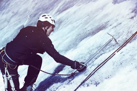 A young man traveler uses an ice ax while hiking in the mountains, a hiker with climbing equipment, in a helmet, crampons climbs to the top of a snow-capped mountain.