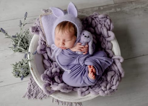 Newborn baby girl wearing knitted hat with rabbit ears holding bunny toy and looking at camera. Infant child kid lying swaddled in purple fabric studio portrait