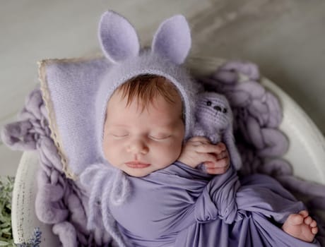 Newborn baby girl wearing knitted hat with rabbit ears holding bunny toy and looking at camera. Infant child kid lying swaddled in purple fabric studio portrait