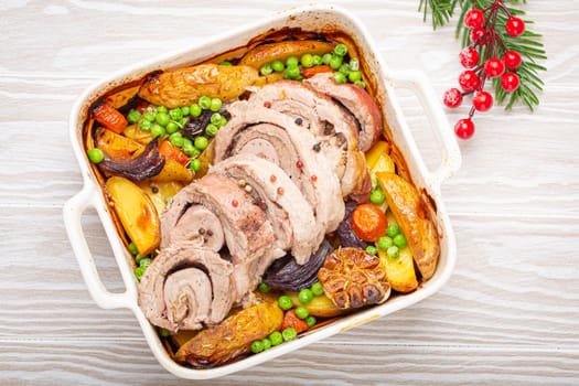 Festive Christmas rolled sliced pork roasted in white casserole dish with potatoes, vegetables and herbs on rustic white wooden background top view. Baked pork roll with vegetables for Xmas dinner.