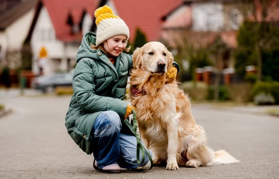 Preteen child girl sitting with golden retriever dog at autumn city street wearing hat and warm jacket. Pretty kid hugging purebred pet doggy labrador outdoors