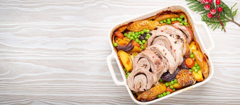 Festive Christmas rolled sliced pork roasted in white casserole dish with potatoes, vegetables and herbs on rustic white wooden background top view. Baked pork roll with vegetables for Xmas dinner.