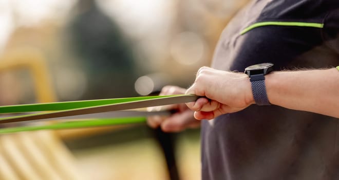 Strong man guy training with elastic rubber band at the stadium outdoors. Adult male person during workout for arms and chest with additional sport equipmant. Sportsman exercising outside
