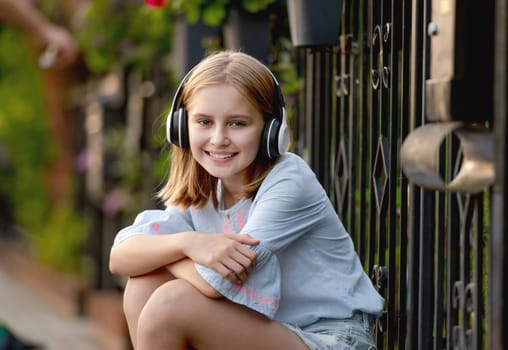 Pretty preteen child girl wearing headphones sitting near fence at city street and smiling. Cute female model with music earpods summer portrait