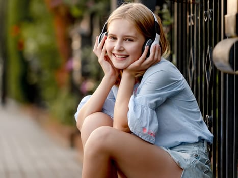 Pretty preteen child girl wearing headphones sitting near fence at city street and smiling. Cute female model with music earpods summer portrait