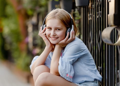 Pretty preteen child girl wearing headphones sitting near fence at city street and smiling. Cute female model with music earpods summer portrait