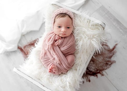 Adorable newborn baby girl lying in tiny bed on fur portrait. Cute infant child kid swaddled in fabric resting in room with daylight