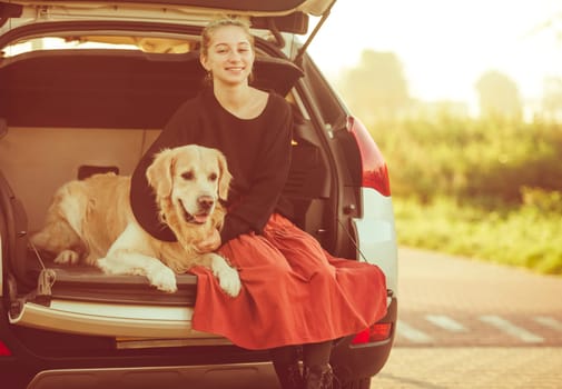 Beautiful girl with golden retriever dog sitting in car trunk and smiling. Pretty teenager hugging purebred pet doggy in vehicle at autumn nature