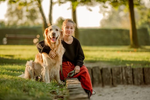 Pretty girl with golden retriever dog sitting at nature and smiling. Beautiful female model teenager hugging purebred pet doggy at autumn park