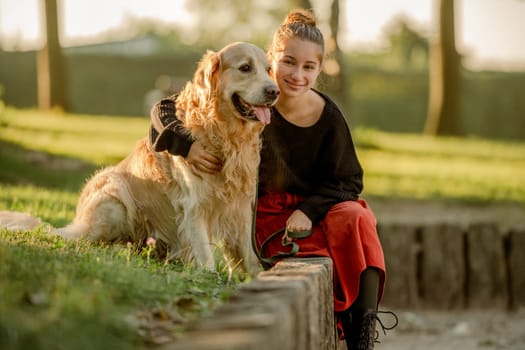 Pretty girl with golden retriever dog sitting at nature and smiling. Beautiful female model teenager hugging purebred pet doggy at autumn park