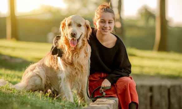 Pretty girl with golden retriever dog sitting at nature and smiling. Beautiful female model teenager hugging purebred pet doggy at autumn park