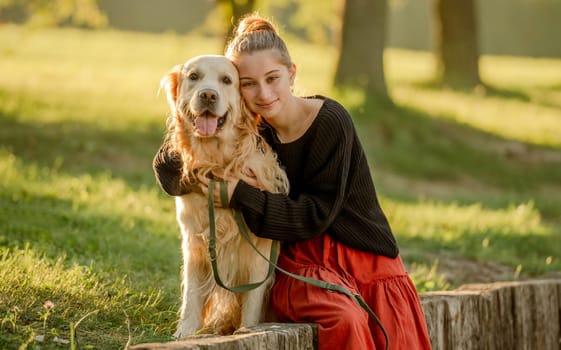 Pretty girl with golden retriever dog sitting at nature and smiling. Beautiful female model teenager hugging purebred pet doggy at autumn park