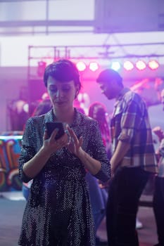 Woman typing message on smartphone while standing on dancefloor in nightclub. Young girl clubber holding mobile phone and texting online in social media while relaxing in club