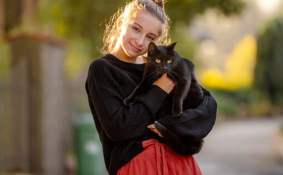 Pretty girl hugging black cat outdoors at street with autumn light. Beautiful model teenager holding feline animal at park