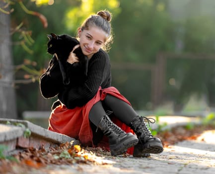 Pretty girl in red skirt hugging black cat outdoors at street with autumn leaves. Beautiful model teenager sitting with feline animal at park