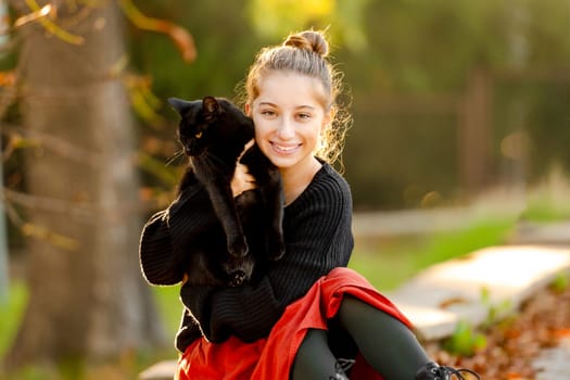 Pretty girl in red skirt hugging black cat outdoors at street with autumn leaves. Beautiful model teenager sitting with feline animal at park