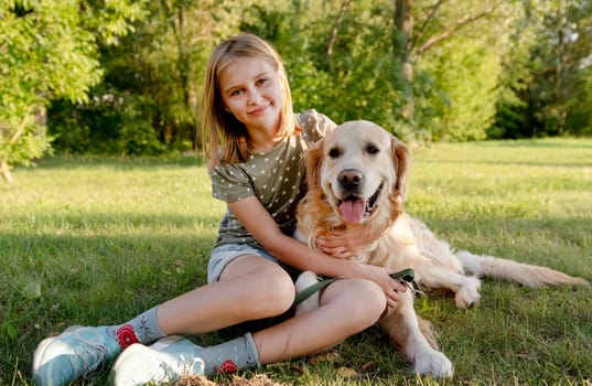 Cute preteen child girl hugging golden retriever dog at nature and sitting at grass. Pretty kid with purebred doggy pet labrador at park