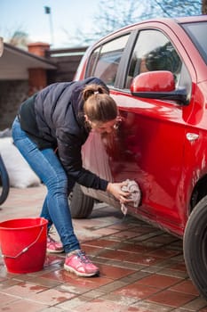 Young slim woman washing red car door with rug