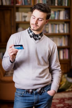 Attractive young man standing at home holding credit card, looking at it