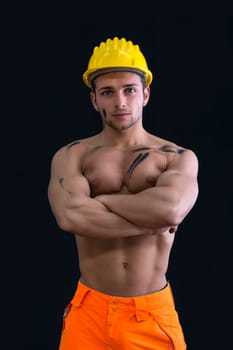 Muscular young construction worker shirtless wearing hardhat, arms crossed, isolated on black background