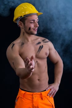 Muscular young construction worker shirtless wearing hardhat, dark background with smoke around