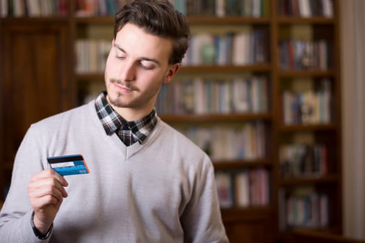 Attractive young man at home holding credit card, looking at it. Large copyspace