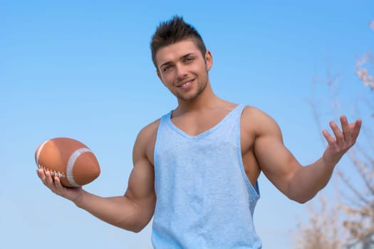 Muscular american football player standing with ball in hand, arms open smiling. Outdoors shot
