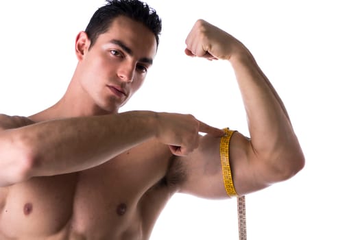 Muscular shirtless young man measuring arm (bicep and tricep) with tape measure, looking at camera, isolated on white