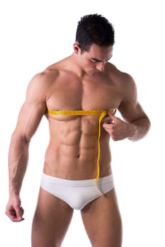Muscular shirtless young man measuring chest and pecs with tape measure, looking at torso, isolated on white