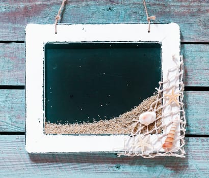 Vintage white wooden frame enclosing a blank black slate with copyspace decorated in one corner with fishing net and seashells on rustic wooden boards, in square format