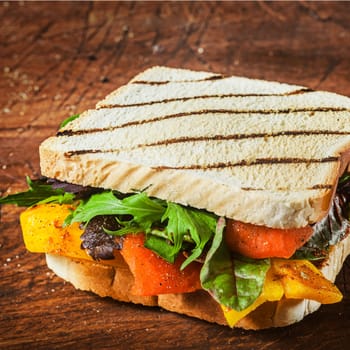 Tasty grilled herb and cheese sandwich on two slices of bread with rocket and spinach, pepper and tomato on a rustic wooden table for a healthy lunchtime snack , close up view