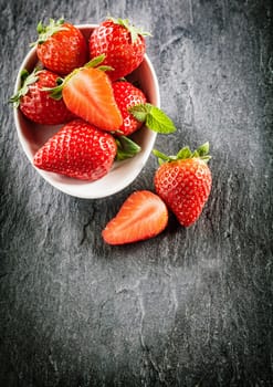 Serving of sweet succulent strawberries in a white bowl garnished with mint with a halved fruit on the slate surface alongside, overhead view with copyspace