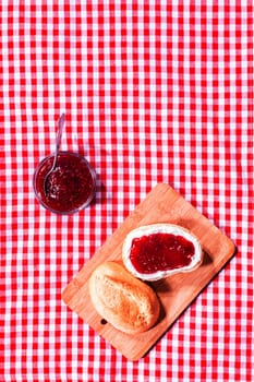 Fresh crusty roll with strawberry jam on a wooden bread board on a red and white checked rustic tablecloth with copyspace, overhead view