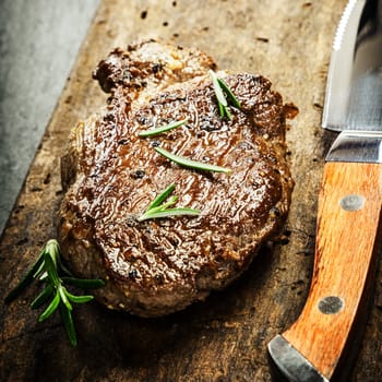 Succulent portion of grilled beef steak garnished with fresh rosemary on a rustic wooden chopping board with a sharp steak knife for a gourmet meal