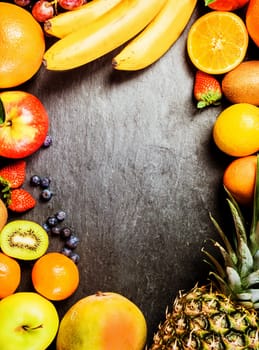 Frame of fresh colorful tropical fruit on grey slate with central copyspace including bananas, orange, pineapple, peach, kiwifruit, apple, strawberries and blueberries