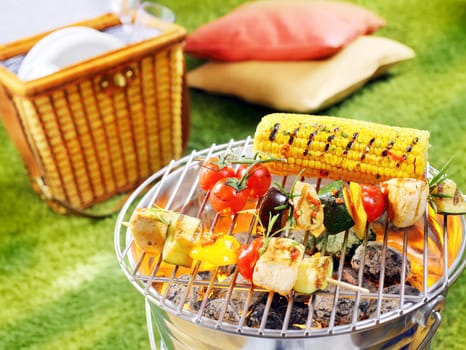 Healthy halloumi kebabs grilling over a fire in a portable barbecue with tomatoes and fresh corn on the cob on a summer picnic in the garden