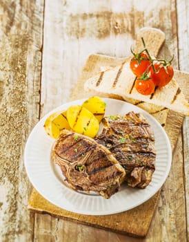 Tasty barbecued picnic lunch with grilled chops and potato with roast cherry tomato and slices of toast served on an old weathered wooden table