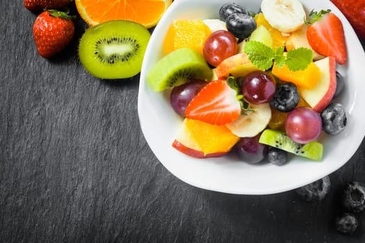 Preparing a bowl of tasty fresh fruit salad for breakfast with tropical ingredients on a rustic slate kitchen counter, overhead view with copyspace