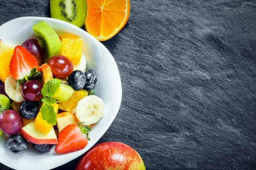 Overhead view of a bowl of fresh fruit salad for a healthy diet made with assorted tropical fruits and an apple, kiwifruit and orange arranged alongside on textured slate with copyspace