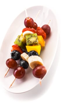 Serving of colorful healthy tropical fruit kebabs on wooden skewers with grapes, banana, orange, kiwifruit and strawberry on an oval white dish, high angle close up view